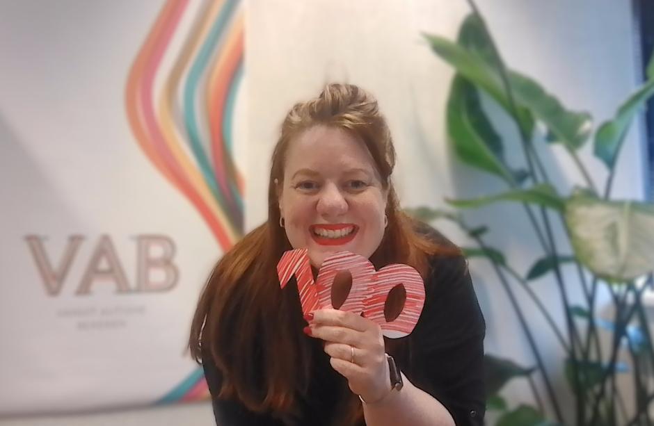 Directeur Anke Laterveer voor een poster van Vanuit autisme bekeken met in haar hand rode uitgeknipte cijfer 100